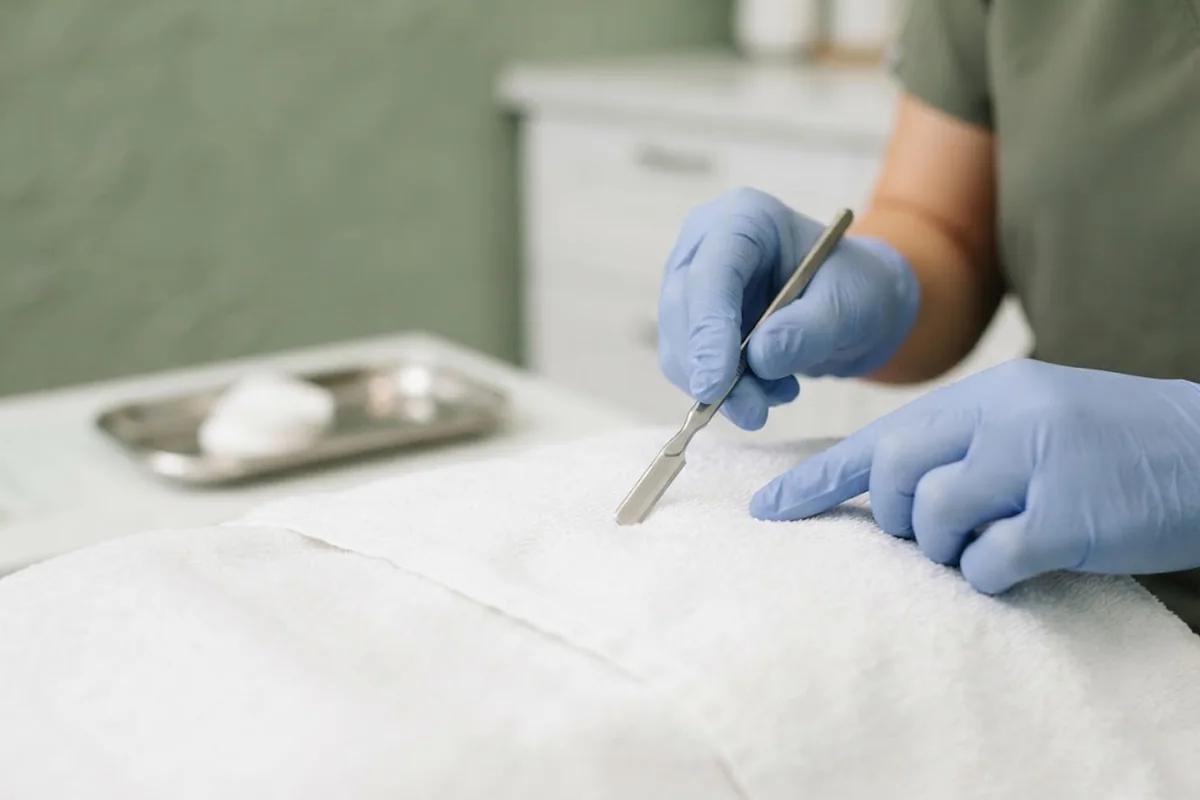 Gloved hand holding a sterile dermaplaning blade on a handle above a white towel in an Irvine CA studio